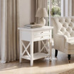 Hampstead 2 Drawer Side Table in white styled beside an upholstered armchair with a ceramic lamp and books, showcasing versatile Hamptons-inspired storage for any room.