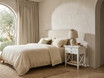 Hampstead 2 Drawer Bedside Side Table in white beside a linen bed with a ceramic lamp and open shelf, creating a relaxed coastal-inspired bedroom setting.
