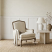 The Cannes Occasional armchair styled with a stone side table and linen throw, featuring a curved French-inspired form, turned legs, and natural linen-blend upholstery