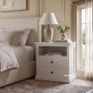 Luxury white nightstand styled with a ceramic lamp and greenery next to an upholstered bed.