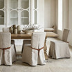 Carlyle Dining Chair with tan leather straps, linen slipcover, and tailored skirt in a neutral modern dining room setting.
