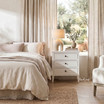 Lando 3 Drawer Bedside Table in white styled beside a cream upholstered bed with a ceramic lamp, books and neutral bedding in a contemporary Hamptons bedroom.
