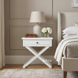 Close-up of the Hampstead Cross Leg Bedside Side Table in white with spacious drawer, cross-leg base, ceramic lamp and decorative accessories in a Hamptons-style bedroom.
