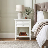 Close-up of the white Hampstead 2 Drawer Bedside Side Table featuring two drawers, open shelf storage and classic Hamptons styling beside a contemporary bed.