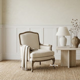 The Cannes Occasional armchair styled with a stone side table and linen throw, featuring a curved French-inspired form, turned legs, and natural linen-blend upholstery