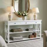 Hampstead 3 Drawer Console with Shelf in white styled beneath a round mirror with matching table lamps, fresh flowers and folded linen in a Hamptons-inspired bedroom.