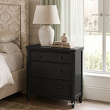 Small black bedside table with three drawers and tapered legs in a styled bedroom with natural light