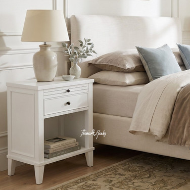 Lando 1 Drawer Bedside Table in white beside an upholstered bed in a Hamptons style bedroom, featuring an open shelf, drawer storage and elegant tapered legs.