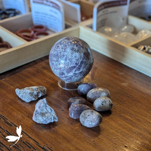 Lepidolite sphere in soft lavender tones displayed with tumbled and raw stones on a wooden table at Prana Blessings Metaphysical Shop in Santa Fe — peace stone known to relieve exhaustion and promote calm. Lepidolite sphere in soft lavender tones displayed with tumbled and raw stones on a wooden table at Prana Blessings Metaphysical Shop in Santa Fe — peace stone known to relieve exhaustion and promote calm.