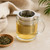 Stainless Steel Tea Cup Infuser - Double Handle resting across a clear glass mug, with its fine micro-hole basket holding loose tea over a pale golden brew.