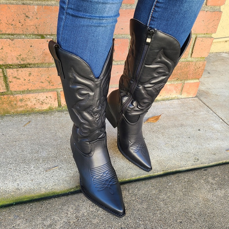 Stylish black cowgirl boots adorned with intricate detailing, the boots are sleek, with pointed toes and a mid-siezed heels, paired effortlessly with blue jeans, captures modern fashion aesthetics while offering a glimpse of urban style. Shop Now!
