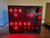 Illuminated control panel with red backlighting, featuring a series of circular buttons in two columns, toggle switches, and a red safety-covered button on the right, against a carbon fiber textured background. The ambient red lighting gives it an intense, high-tech appearance, suggestive of gaming or simulation gear