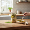 Pickled asparagus jar on a wooden kitchen counter with fresh garlic, dill, and a hand serving asparagus spears into a bowl.