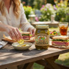 Million Dollar Pickles jar on a picnic table with ham sandwiches, a fresh salad, and iced tea in a sunny outdoor setting.
