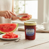 A jar of Harvest Array Watermelon Preserves sits on a light wooden table beside a plate of fresh watermelon cubes and a large watermelon slice.