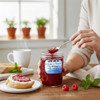 A jar of Harvest Array Red Raspberry Preserves sits on a light wooden table with fresh raspberries beside it. In the background, a person spreads the vibrant red preserves onto a slice of bread with cream cheese.
