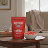 A bright red bag of Old School Brand Cinnamon Puffs next to a bowl of the Cinnamon Puffs on an end table next to  a  cozy sofa in a living room. Make a great treat for company or movie night.