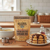 A country kitchen on a bright morning. A 16 oz. package of Old School Brand Chocolate Chip Pancake Mix is on a wooden counter top between a  mixing bowl and a plate of freshly made chocolate chip pancakes.