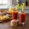 Two Bloody Mary's with J.Q. Dickinson Salt Works Bloody Mary Salt around the rims and celery sticks as garnish, on a dark kitchen counter with a container of Bloody Mary Salt. and brunch items.