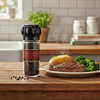 A bright country kitchen setting with a dinner plate of steak, baked potato and broccoli next to a reusable pepper grinder with Tellicherry Peppercorns on a wood kitchen counter.