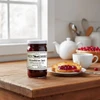 Annie's Kitchen Cranberry Jam Jar next to a plate of toast with cranberry jam on it, a tea cup , teapot, and basket of cranberries on a wooden table in a bright kitchen.