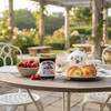 A lovely outdoor tea party with a jar of  Mrs. Miller's Homemade Bumbleberry Jam, a plate of croissants, a bowl of strawberries, raspberries, and blueberries on a garden patio table. A white teapot and cups with a strawberry motif are also on the table.