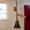 Oak wood Amish-Made Wall-Mount Broom Holder fixed to a kitchen wall with someone holding a Stoltzfus Broom next to it, getting ready to put it in the holder.