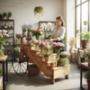 Woman arranging flowers on a wooden tiered display cart with black metal wheels inside a bright flower shop