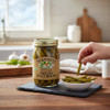 Pickled dilly beans jar on a wooden kitchen counter with garlic, fresh dill, and a hand serving green beans into a small bowl.