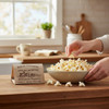 Hand reaching into a bowl of Amish Country white hulless butter microwave popcorn on a kitchen counter beside the boxed package.