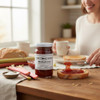 Annie’s Kitchen Rhubarb Jam being spread on toast with fresh rhubarb stalks, bread, and coffee on a wooden table.
