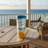 Welcome to the Beach 16 ounce Tumbler with a blue Lid on an wooden patio table table with a view of the ocean in the background.
