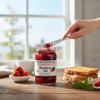 No-sugar-added strawberry jam jar on a wooden table with fresh strawberries, toast being spread, and a warm drink in the background.