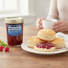 A jar of Harvest Array Raspberries & Brandy preserves sits on a rustic wooden table, surrounded by fresh raspberries, a steaming cup of tea, and a buttermilk biscuit.