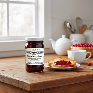 Annie's Kitchen Cranberry Jam Jar next to a plate of toast with cranberry jam on it, a tea cup , teapot, and basket of cranberries on a wooden table in a bright kitchen.
