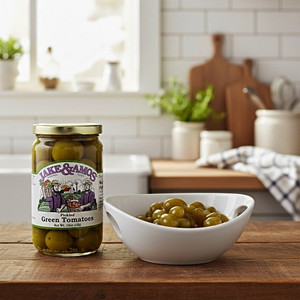 A jar of  Jake & Amos Pickled Green Tomatoes next to a bowl of the green tomatoes on a wooden kitchen counter in a country style kitchen.