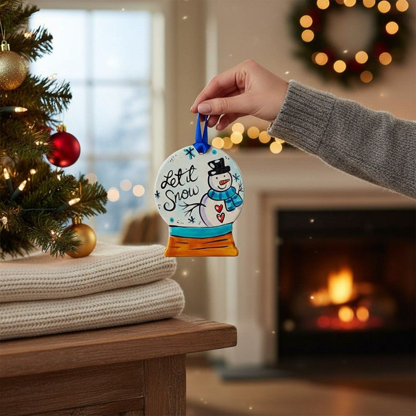 Hand-painted let it snow snowman ornament being hung on a Christmas tree with warm holiday lights in the background