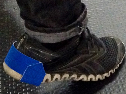 Person's ankle and foot in jeans and a sneaker with a blue heel grounder attached under the heel and around the back of the shoe.