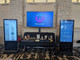 Two 55-inch 500nit 24x7 touch screen digital poster kiosks flanking a table at a professional trade show display.
