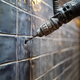 A power drill using a bit from the Add Tile Wall Installation Kit to create a hole in a dark, textured tile wall.
