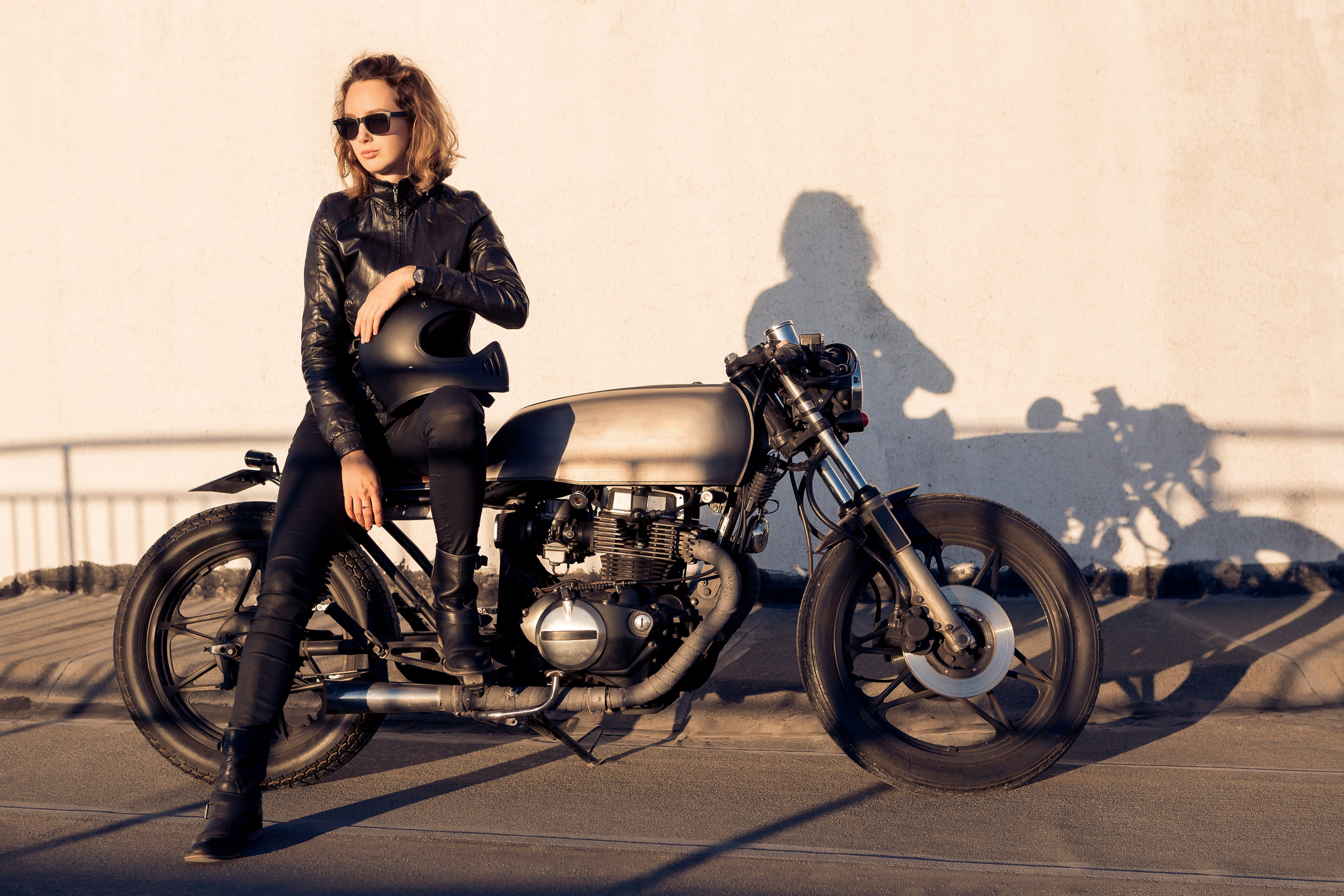 Women leaning on a motorcycle with her shadow reflecting against the wall behind her. 