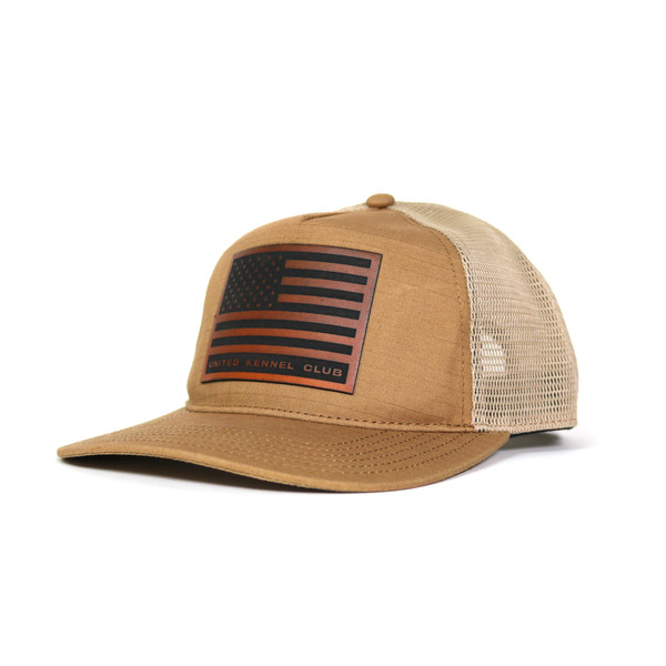 A tan trucker-style snapback hat featuring a structured canvas front and white mesh back. The front panel displays a rectangular faux leather patch engraved with the American flag and United Kennel Club logo.