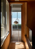 A hallway showcases a polished chrome Flowerpot VP12 floor lamp with a beige shade and white marble base, standing on brown carpet. Windows are on the left, a wooden banister on the right, and an open doorway ahead reveals a bright room with a water view.