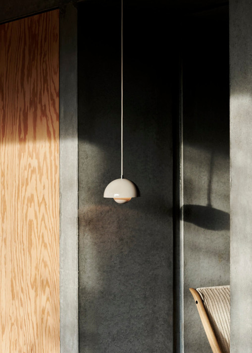 A neutral-toned Flowerpot VP10 pendant lamp hangs from a light-colored cord against a dark gray wall, partially lit by sunlight. To the left, a light wood panel wall is visible.
