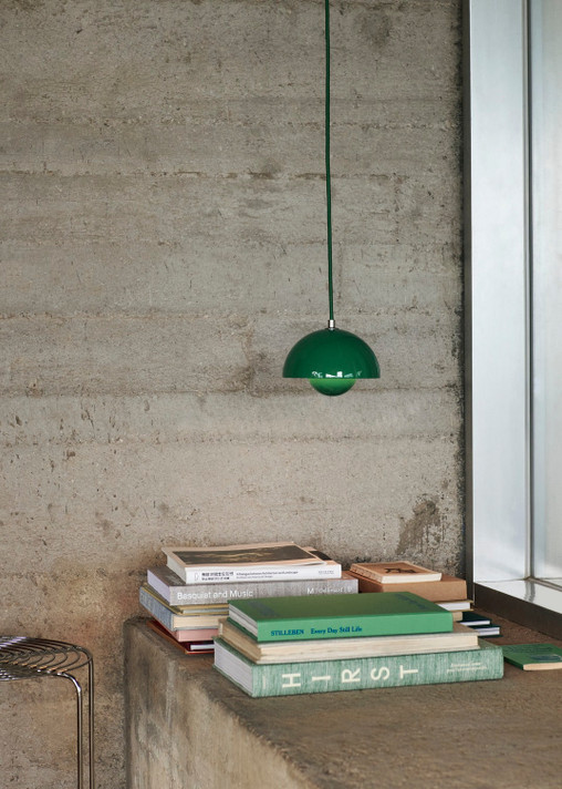 A signal green Flowerpot VP10 pendant lamps hang over a textured concrete wall. Below, stacks of books, rest on a concrete ledge and a metal wire stand.