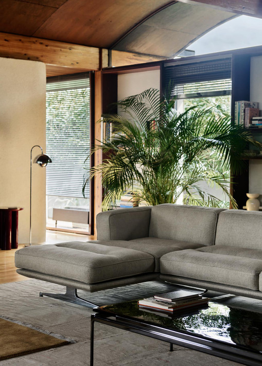A warm living space features a gray L-shaped sectional sofa with a dark coffee table. A polished chrome Flowerpot VP12 floor lamp stands left of the sofa, with large windows, a large green plant, and wooden ceilings in the background.