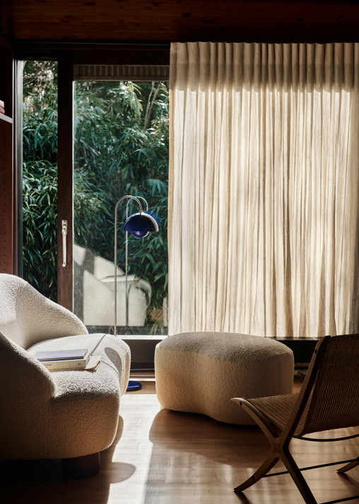 A modern living room features a creamy boucle lounge chair and ottoman, a wooden chair, and a cobalt blue Flowerpot VP12 floor lamp by large windows with sheer curtains and a view of lush greenery.