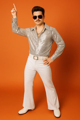 Fantasy Costumes Shirt Disco- Dazzling Sequin Shirt, a man wearing a silver sequin disco shirt, white wide-leg pants, and sunglasses, posing against an orange background with one arm raised.