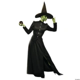 Incharacter Classic Witch Costume Deluxe Adult, a woman dressed as a witch with green skin, wearing a tall black pointed hat, a fitted black dress with silver buttons, and a long black cape, holding a crystal ball in one hand and adjusting her hat with the other.