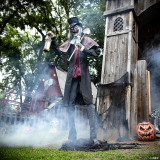 Seasonal Visions 8' 6'' Animated Graveyard Host, a life-sized sinister skeleton in a dark Victorian-style outfit holding a lantern and shovel, standing in a spooky graveyard scene with fog, tombstones, and Halloween decorations.